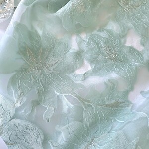 May include: Close-up of a delicate, sheer fabric in a soft, pale green hue. The fabric is adorned with an intricate floral pattern, featuring raised, textured flowers with silver accents. The design creates a sense of depth and elegance, suitable for apparel or home decor.