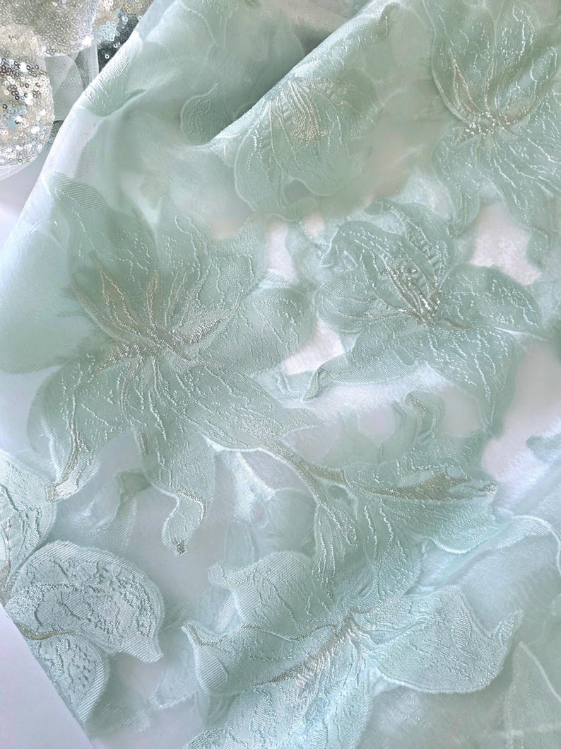 May include: Close-up of a delicate, sheer fabric in a soft, pale green hue. The fabric is adorned with an intricate floral pattern, featuring raised, textured flowers with silver accents. The design creates a sense of depth and elegance, suitable for apparel or home decor.