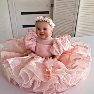 May include: A baby doll wearing a pink dress with floral accents and a matching floral headband. The dress features a layered skirt and puffy sleeves. The text "fedchyshyna_babydress" is visible on the dress.
