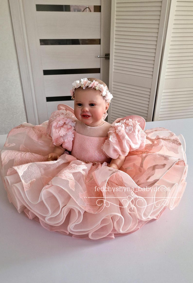 May include: A baby doll wearing a pink dress with floral accents and a matching floral headband. The dress features a layered skirt and puffy sleeves. The text "fedchyshyna_babydress" is visible on the dress.