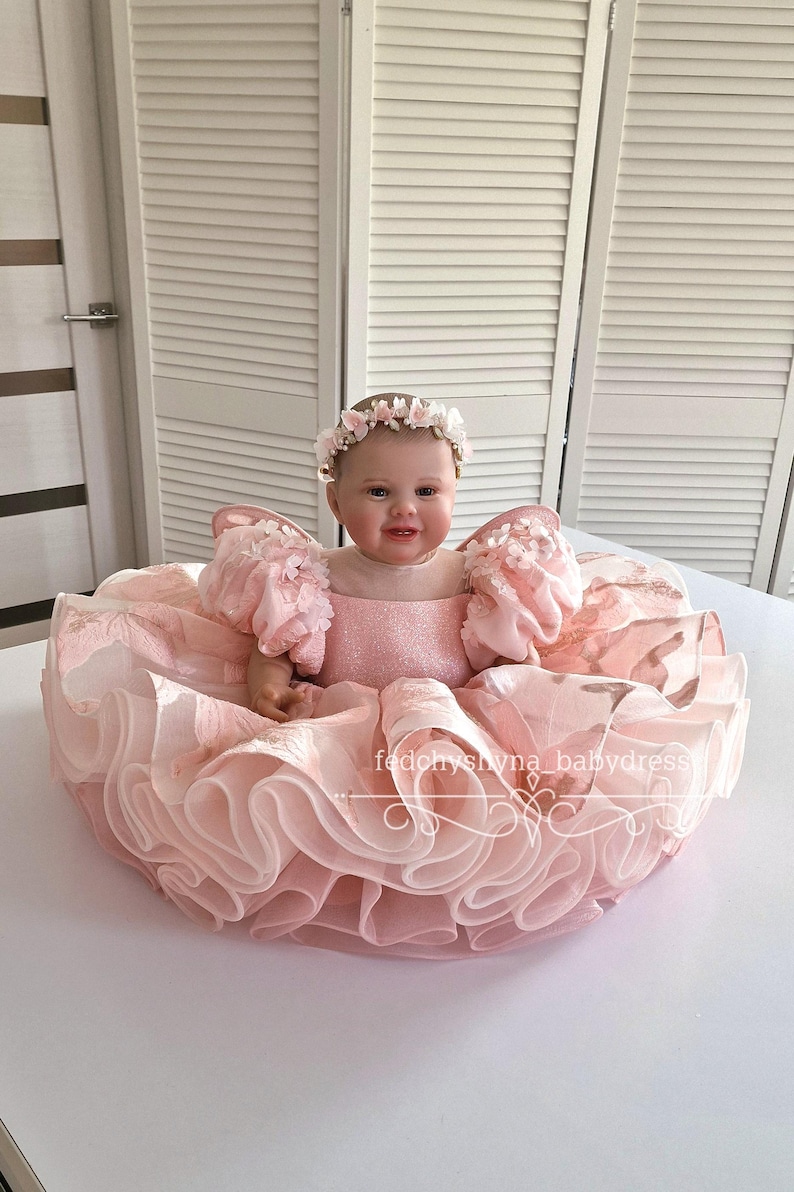 May include: A baby doll wearing a pink dress with ruffled layers and puffy sleeves. The doll has a floral headband and is seated in a round, ruffled dress. The dress has the text "fedonyshyna babydress".
