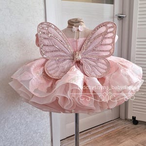 May include: A pink dress with a layered skirt and a butterfly wing design. The dress is displayed on a stand. The butterfly wings are embellished with glitter. The dress has a tulle skirt and a bodice with floral details.