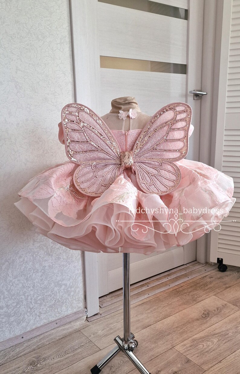 May include: A pink dress with a layered skirt and a butterfly wing design. The dress is displayed on a stand. The butterfly wings are embellished with glitter. The dress has a tulle skirt and a bodice with floral details.