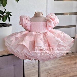 May include: A pink baby dress with a layered skirt and puffy sleeves. The bodice is embellished with glitter and the sleeves have floral accents. The dress is displayed on a metal stand. The text "fedchyshyna_babydress" is visible.