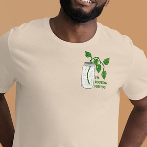 Puede incluir: Una persona lleva una camiseta beige claro con una planta verde en un frasco de vidrio que dice "Te estoy animando".
