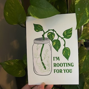 以下が含まれることがあります： ガラスの瓶に入れた緑色の植物が描かれた白いカード。植物は緑と黄色の葉をしています。カードには「応援しています」と書かれています。