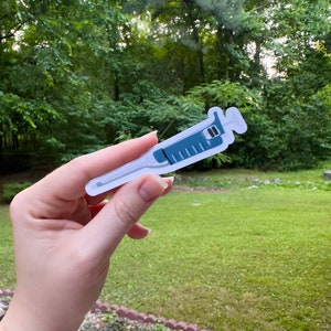 May include: A white sticker with a blue syringe illustration. The syringe has a needle and markings on the side.