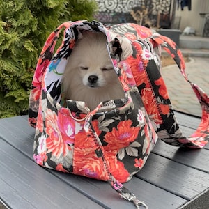 以下が含まれることがあります： 黒と赤のフローラルなペットキャリアに、小さな白い犬が入っています。キャリアはジッパーで閉じることができ、ショルダーストラップが付いています。