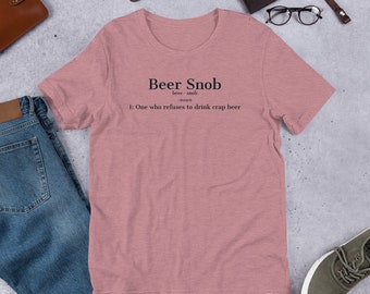 Puede incluir: Una camiseta de manga corta color rosa empolvado con texto negro que dice: Beer Snob, beer · snob, -noun, 1: One who refuses to drink crap beer. La prenda se muestra junto a unos vaqueros azules, una cartera de cuero marrón, un reloj y gafas.