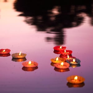Puede incluir: Velas flotantes en varios colores, incluyendo rojo, amarillo y azul, con llamas encendidas, se reflejan en el agua. La superficie del agua es de un suave púrpura, con un reflejo borroso del cielo.