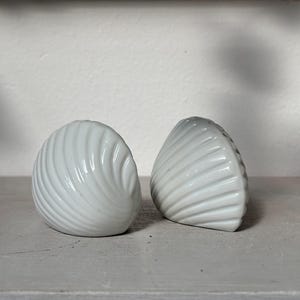 May include: Two white ceramic seashell-shaped salt and pepper shakers. The shakers have a glossy finish and a detailed, ridged design. They are displayed on a light grey surface against a white background.