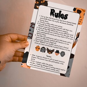 May include: A printed game card with the word "Rules" at the top. The card features instructions for a horror movie-themed bingo game, with illustrations of iconic movie villains. The card is held by a person.
