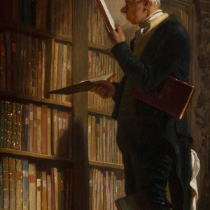 May include: A man in a dark suit and white shirt stands in a library, holding a book in his left hand and looking at another book in his right hand. The library shelves are filled with books.