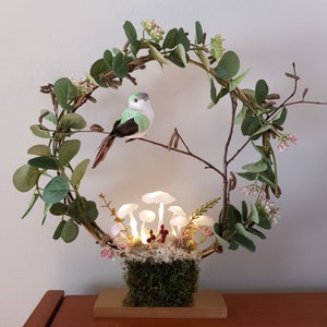 Könnte beinhalten: Eine grüne Vogelfigur, die auf einem Zweigkranz mit einer kleinen moosbewachsenen Basis und weißen Pilzlichtern sitzt. Der Kranz besteht aus Eukalyptusblättern und hat einen goldenen Drahtrahmen.