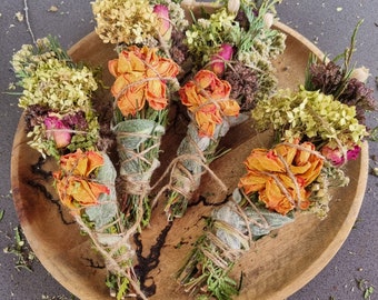 Può includere: Quattro mazzetti di erbe e fiori essiccati disposti su un vassoio rotondo in legno. Ogni mazzetto è legato con dello spago e presenta rose arancioni, ortensie gialle, fogliame verde e piccoli fiori viola, creando una composizione botanica.