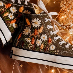 Op de afbeelding: Olijfgroene hoge sneakers met gedetailleerde bloemenborduurwerk. Het ontwerp toont witte madeliefjes, tarwearen en wilde bloemen in oranje, bruin en groen. De sneakers hebben witte veters en een witte rubberen zool met een zwarte streep.
