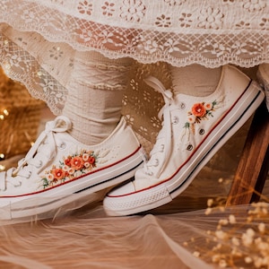 May include: White canvas sneakers with floral embroidery in shades of orange and red. The shoes have a red and black stripe detail on the sole. The laces are white. The sneakers are a low-top style.