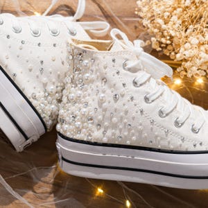 May include: White high-top sneakers embellished with pearls and rhinestones. The shoes have white laces, a white rubber sole with a black stripe, and a canvas upper. The sneakers are decorated with various sizes of pearls and rhinestones.