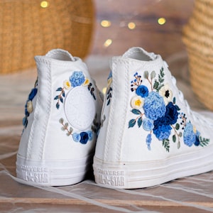 Puede incluir: Zapatillas altas blancas con diseños florales bordados en azul y amarillo. Las zapatillas presentan el logotipo de Converse All Star en el talón y el lateral. El bordado floral incluye varios tonos de azul, amarillo y verde.