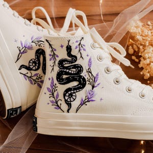 Puede incluir: Zapatillas blancas de caña alta con una serpiente negra y bordado floral morado.