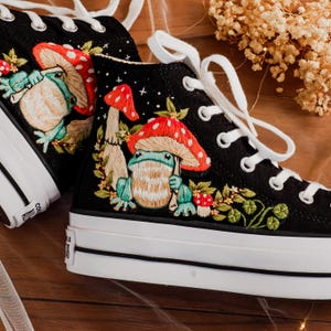 Op de afbeelding: Zwarte hoge canvas schoenen met geborduurde kikkers en paddenstoelen. De schoenen hebben witte veters en een witte zool.