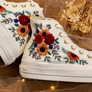 May include: Pair of white high-top sneakers with colorful floral embroidery. The embroidery features sunflowers, red flowers, and blue berries, with green leaves. The sneakers have white laces and eyelets.
