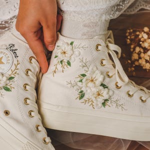 May include: White high-top sneakers with gold eyelets and embroidered white flowers and green leaves. The left shoe has the word "Robert" written on the side.