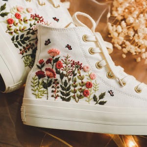 Puede incluir: Zapatillas blancas de lona de caña alta con diseños florales bordados en tonos rosa, rojo y verde. Las flores están dispuestas en un racimo en el lateral del zapato.