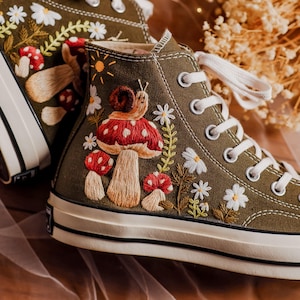 Puede incluir: Zapatillas de lona de caña alta de color verde oliva con champiñones, flores y un caracol bordados. Las zapatillas tienen cordones blancos y una suela de goma blanca.