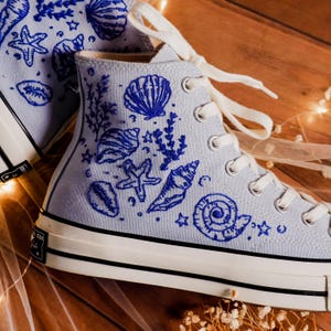 May include: Light blue high-top sneakers with white laces and a black and white sole. The sneakers are embroidered with blue seashells, starfish, and aquatic plants. The shoes are on a wooden surface.