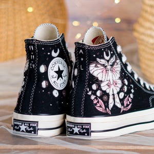 Könnte beinhalten: Schwarze High-Top-Sneaker mit weißen Sohlen und Schnürsenkeln. Die Sneaker haben ein Motiv aus Motten und Monden mit rosa und weißen Akzenten. Das Converse All Star-Logo ist auf der Seite und der Zunge zu sehen.