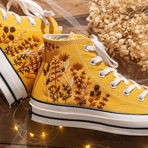 Könnte beinhalten: Senfgelbe High-Top-Sneaker mit gestickten Blumendesigns in Braun-, Beige- und Weißtönen. Die Sneaker haben weiße Schnürsenkel, eine weiße Gummisohle und einen schwarzen Streifen. Eine stilvolle und einzigartige Schuhoption.