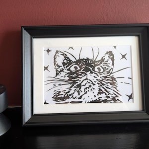 Puede incluir: Un grabado en linóleo en blanco y negro de una cara de gato con ojos grandes y bigotes, rodeado de estrellas de cuatro puntas. La impresión está en un marco negro rectangular con paspartú blanco, junto a un portavelas negro cilíndrico.