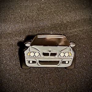 May include: A silver enamel pin of a gray BMW M3 car with black headlights and a black grill.