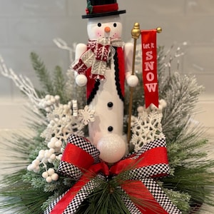 May include: A snowman figurine wearing a black top hat, red vest, and scarf, holding a banner that says "Let it Snow." The snowman is surrounded by decorative greenery, snowflakes, and a red and black checkered bow.