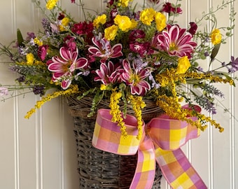 May include: A woven basket overflowing with colorful artificial flowers, including yellow, burgundy, and pink blooms. A large, decorative bow with a pink, yellow, and orange plaid pattern adorns the front of the basket, which is hanging on a white paneled wall.