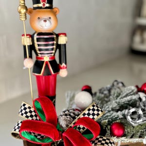 May include: A decorative Christmas bear nutcracker figurine in a soldier's uniform, holding a gold staff. The bear is wearing a black hat with red accents. A red velvet bow with a black and white checkered ribbon is in the foreground.
