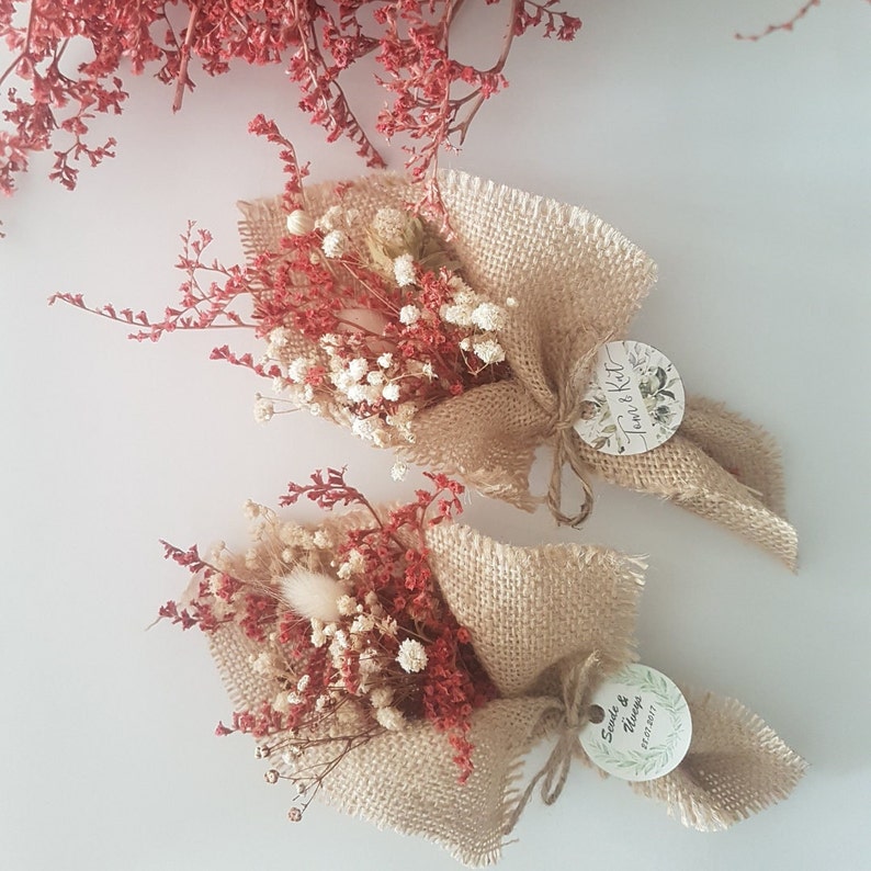 May include: Two small bouquets of dried flowers wrapped in burlap with a personalized tag attached to each. The tags are round and feature a white background with black text. The top tag says "Tom & Kat" and the bottom tag says "Sarah & Thomas 21.07.2017".