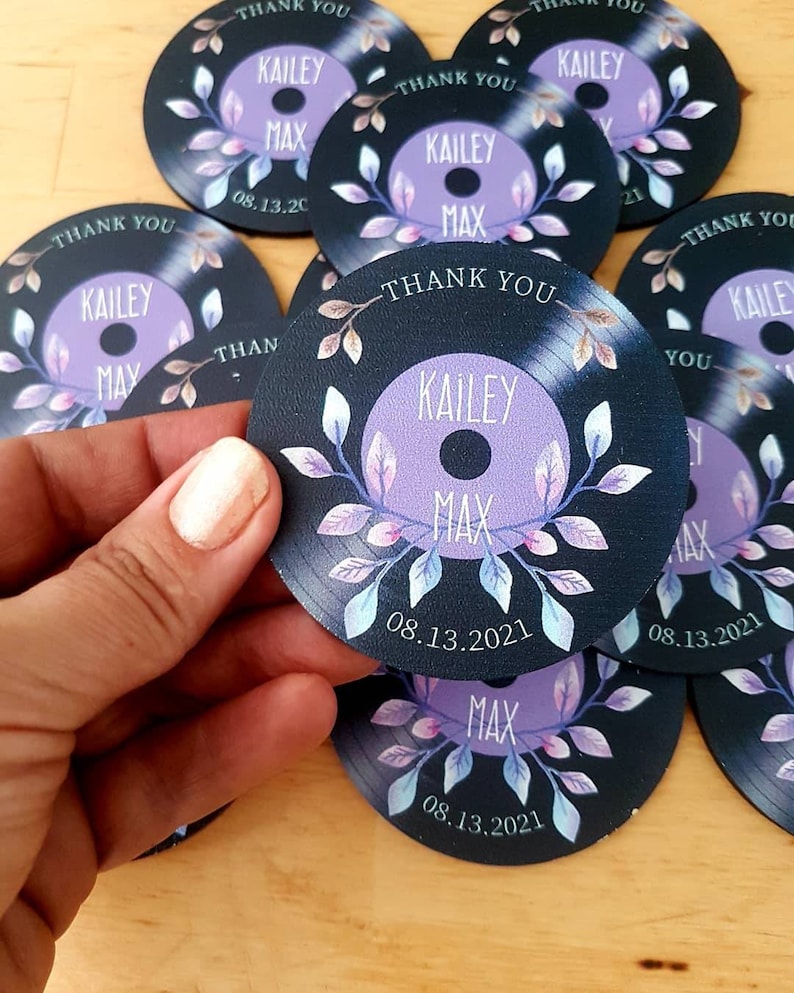 May include: Circular thank you favors with a black and white vinyl record design. The record has a purple center with the names "Kailey" and "Max" and the date "08.13.2021". The record is surrounded by a floral design with leaves and flowers in shades of pink and blue.