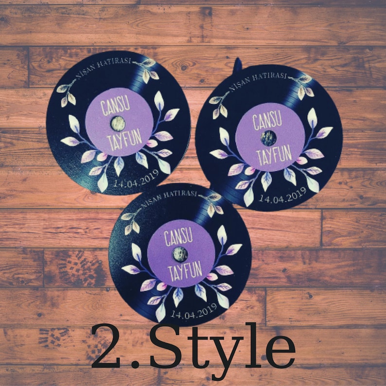 May include: Three black vinyl record-shaped wedding favors with purple and white floral designs. The text "NISAN HATIRASI" is printed on each record, along with the names "CANSU" and "TAYFUN" and the date "14.04.2019".