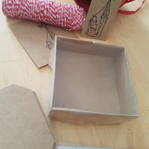 May include: A small, empty, brown cardboard box with a clear plastic lid. The box is sitting on a wooden surface. There is a roll of red and white twine, a brown tag with a drawing of a person and the text "@littleventuresplus", and a brown paper envelope in the background.
