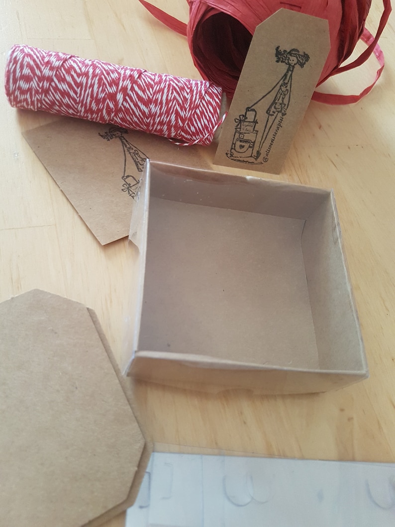May include: A small, empty, brown cardboard box with a clear plastic lid. The box is sitting on a wooden surface. There is a roll of red and white twine, a brown tag with a drawing of a person and the text "@littleventuresplus", and a brown paper envelope in the background.