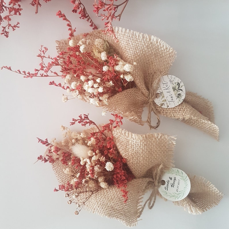 May include: Two small bouquets of dried flowers wrapped in burlap with a circular tag attached to each. The tag on the left bouquet says "Tom & Kat" and the tag on the right bouquet says "Sande & Nanya 21.07.2017".