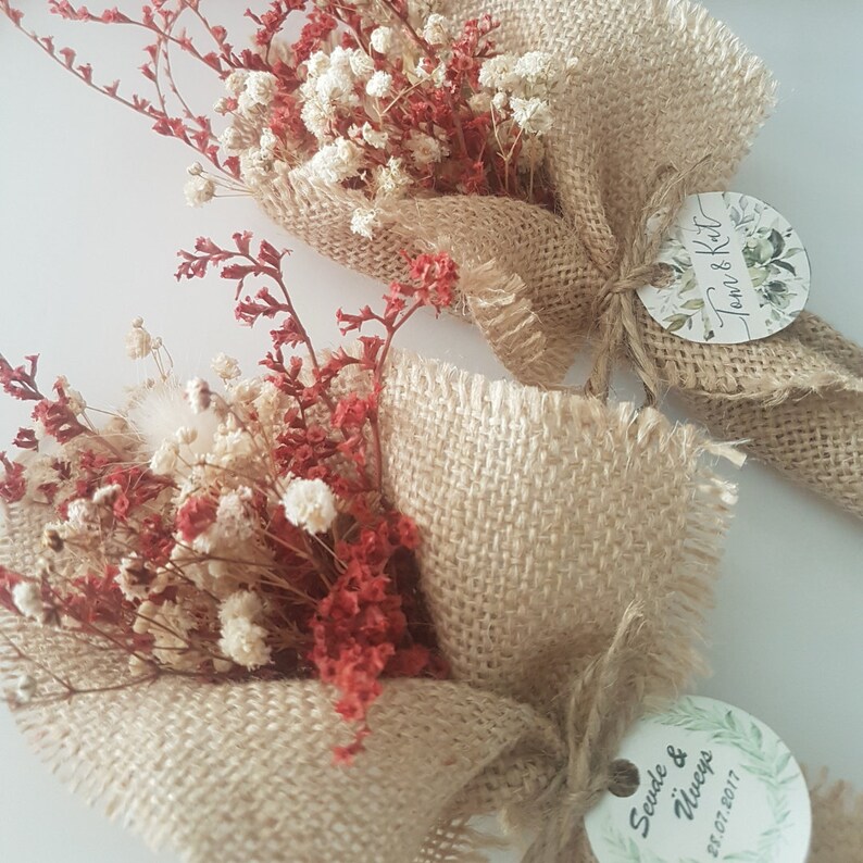 May include: Two small bouquets of dried flowers wrapped in burlap fabric. The bouquets are tied with twine and have a small round tag attached. The tag on the left bouquet says "Sevda & İlker" and the date "21.07.2017". The tag on the right bouquet says "Tom & K&uuml;t" and has a floral design.