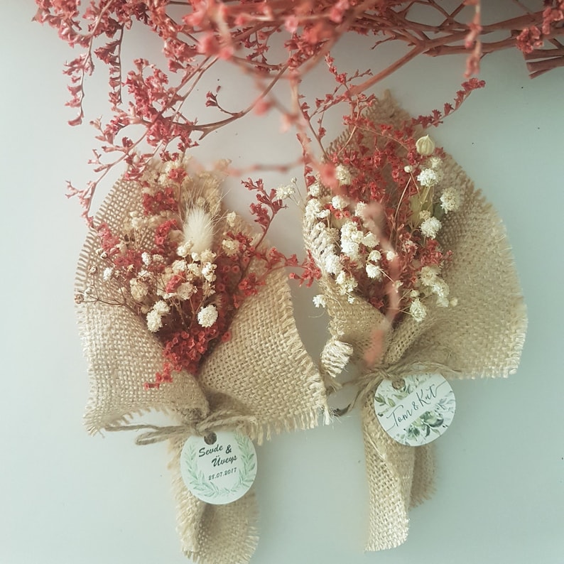 May include: Two small bouquets of dried flowers wrapped in burlap fabric with a circular tag attached to each. The tag on the left bouquet has the text "Sevda & Always 22.07.2017" and the tag on the right bouquet has the text "Tom & Kat 22.07.2017".