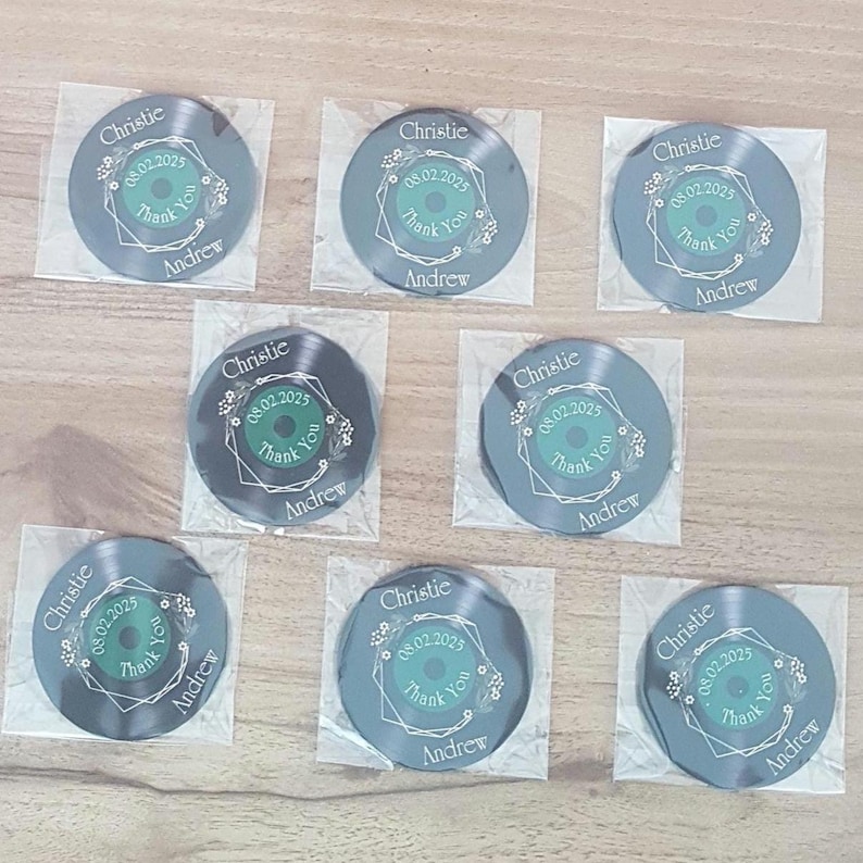 May include: Eight black vinyl record-shaped thank you favors with a green and white floral design. The text on the favors reads "Christie & Andrew 08.02.2025 Thank You".