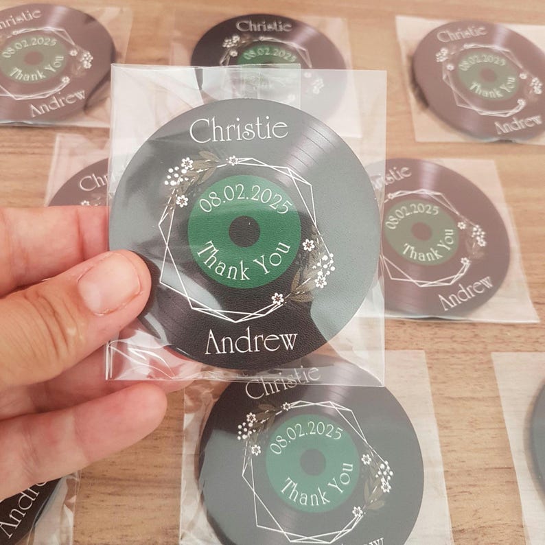 May include: Black and white vinyl record shaped wedding favor with a green circle in the center. The circle has the text "Thank You" and the date "08.02.2025". The record also has the names "Christie" and "Andrew" printed on it.