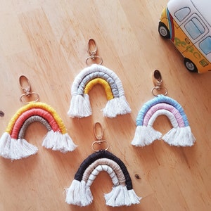 May include: Four rainbow-coloured macrame keychains with white tassels. Each keychain features a different colour combination: orange, yellow, and red; blue, pink, and white; black, beige, and white; and grey, yellow, and white.