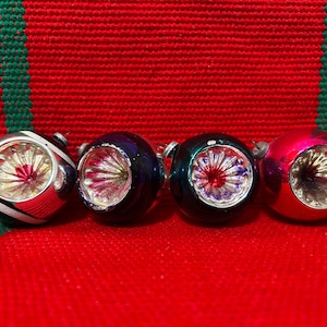 Pode incluir: Quatro enfeites de Natal vintage em fila. Os enfeites são prateados, azul escuro, verde-azulado e rosa. Cada um tem uma base prateada e um centro vermelho ou roxo. Os enfeites estão em um fundo vermelho com listras verdes.