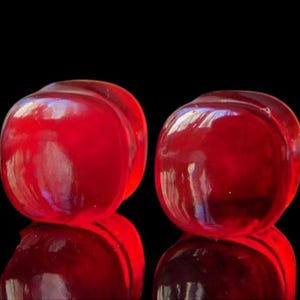 May include: Two translucent, red, rounded objects with a glossy finish are set against a black background. The objects are reflected below, creating a symmetrical composition. The shapes appear to be smooth and solid.